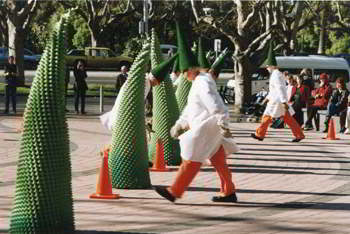 Actors in white coats and cone hats move amongst freestanding tall green cones